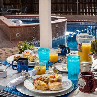 A patio table at Woodrow House Bed & Breakfast is set for breakfast with plates of food, blue tumblers, and fresh juice. In the background, a hot tub and pool with water jets create a relaxing B&B near Texas Tech University atmosphere.