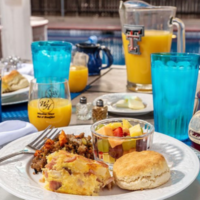 A plate with a biscuit, egg casserole, hash brown casserole, and fruit sits on a table next to blue glasses and orange juice at Woodrow House Bed & Breakfast—a charming B&B near Texas Tech University, with a pool in the background.