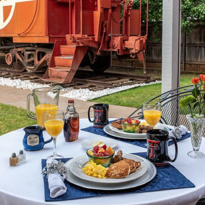 A table set for breakfast outdoors at Woodrow House Bed & Breakfast features scrambled eggs, pancakes, fruit, mugs, orange juice, and syrup. In the background, a red train car sits near a grassy yard—perfect Texas Tech lodging.