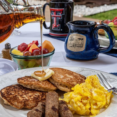 A breakfast plate at Woodrow House Bed & Breakfast features scrambled eggs, sausage links, and pancakes with butter and syrup. A bowl of mixed fruit and mugs sit in the background—a cozy B&B near Texas Tech University.