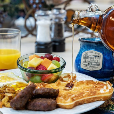 A breakfast plate at Woodrow House Bed & Breakfast features waffles with syrup, sausage links, potatoes, fried eggs, and mixed fruit. Enjoy Texas Tech lodging with fresh orange juice and condiments in the background.