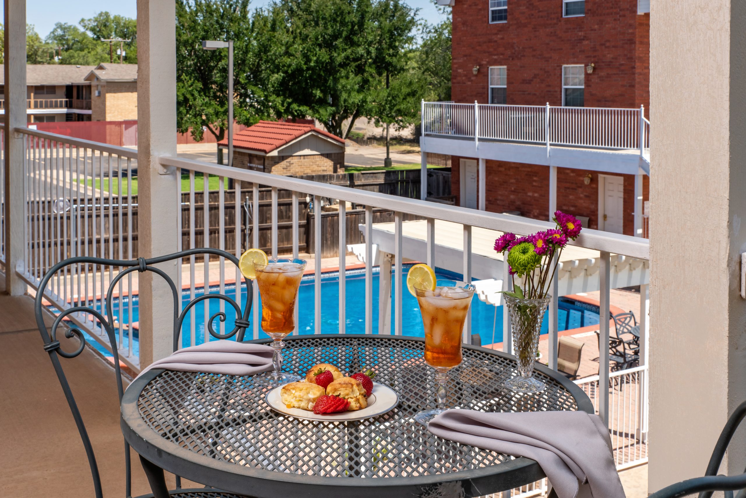 A round metal table at Woodrow House Bed & Breakfast overlooks a pool. Two iced teas with lemon, pastries, napkins, and a vase of flowers await as you relax in this inviting B&B near Texas Tech University, with trees visible in the background.