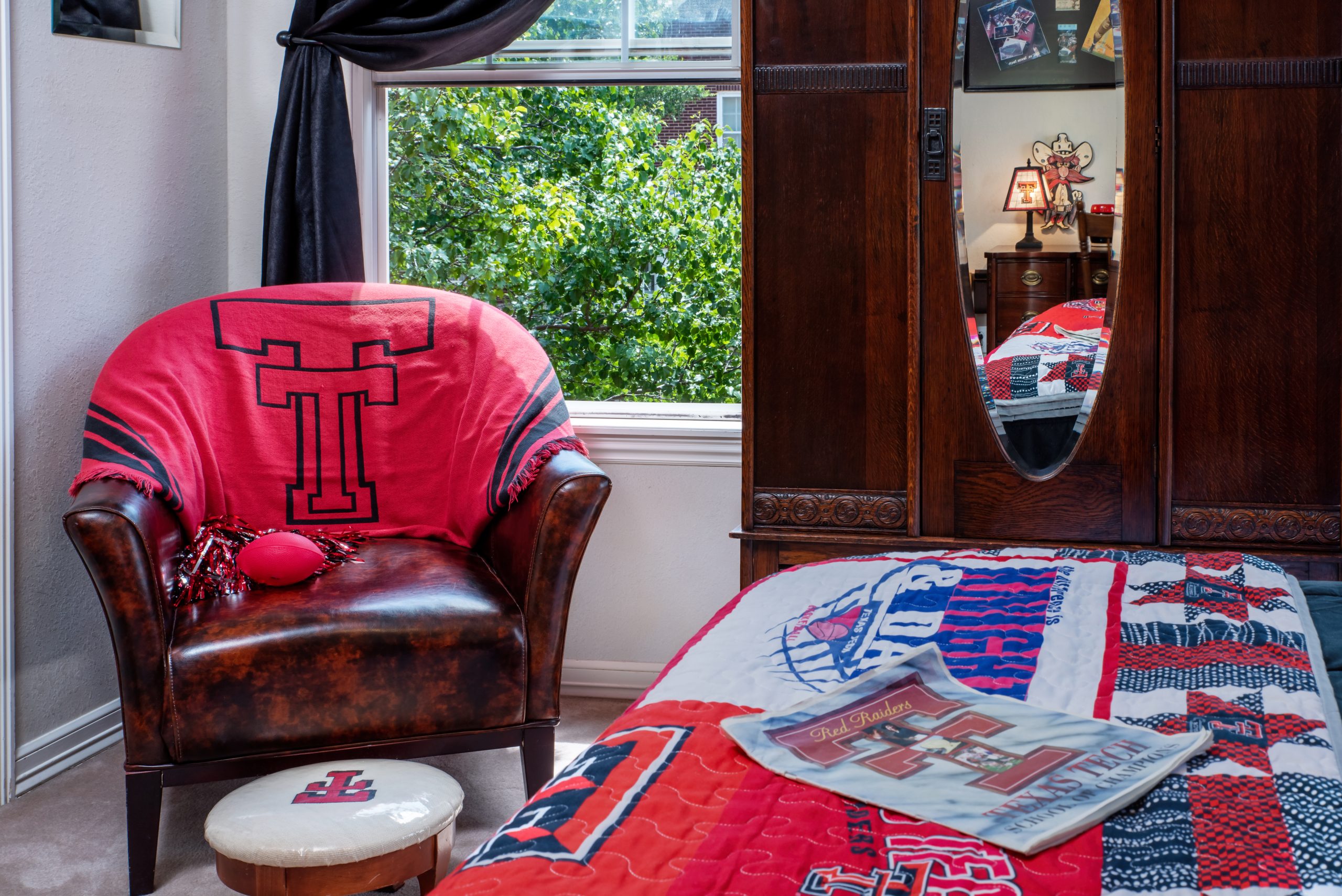 A cozy bedroom at Woodrow House Bed & Breakfast, adorned with Texas Tech University decor—a TT logo quilt, brown leather chair with a red throw, and a magazine on the bed. The window reveals green trees outside, perfect Texas Tech lodging.