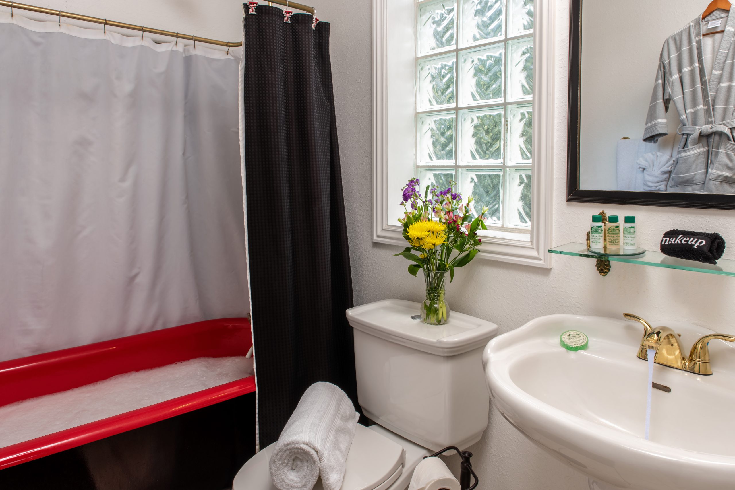 A bathroom at Woodrow House Bed & Breakfast features a red bathtub filled with bubbles, a toilet topped with rolled towels, fresh flowers in a vase, and a white sink with running water—perfect Texas Tech lodging near the university.