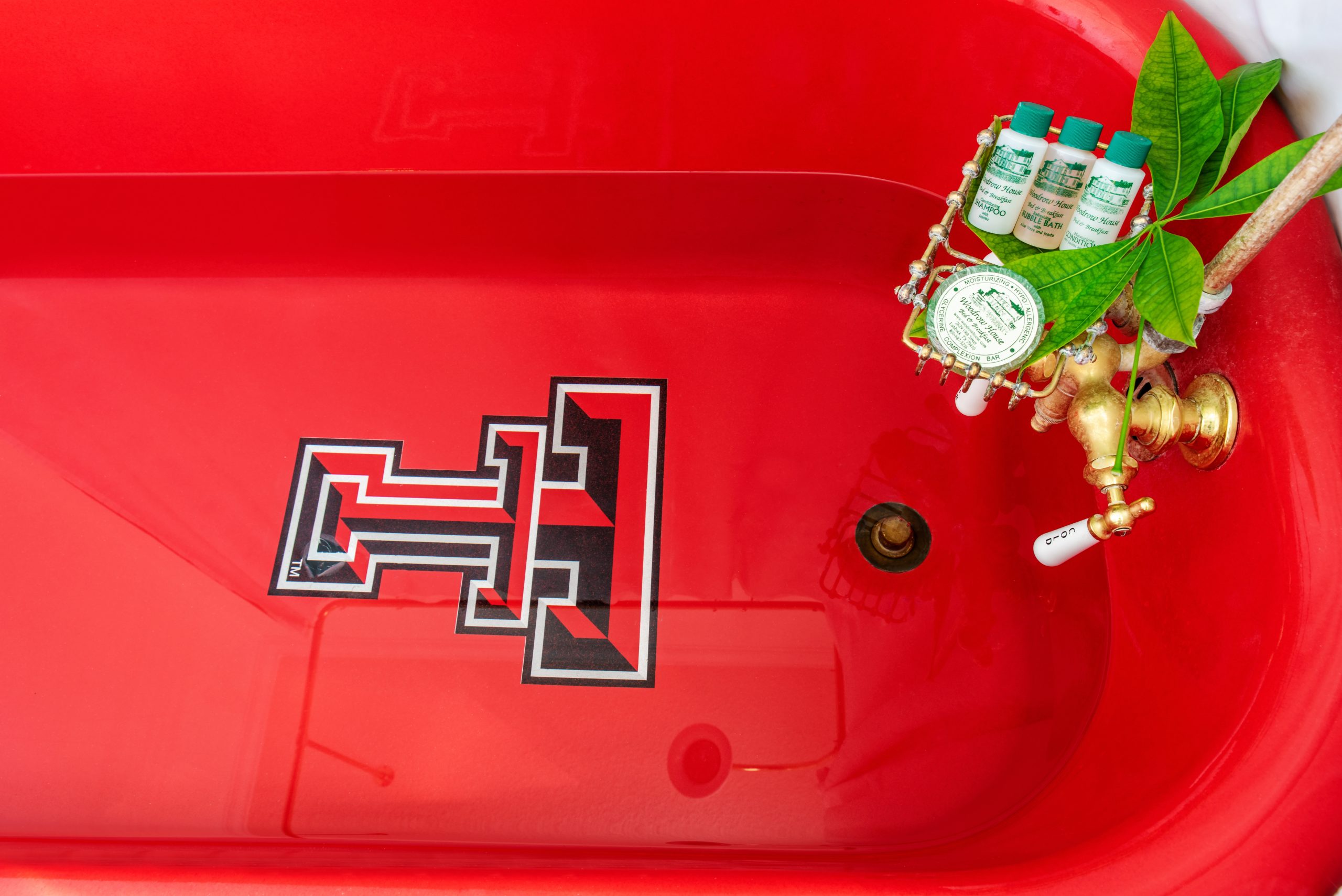 A bright red bathtub features the Texas Tech University double-T logo at the bottom. The gold faucet holds four small bottles, a soap dish, and a leafy green plant—just one unique touch at Woodrow House Bed & Breakfast, premier Texas Tech lodging.