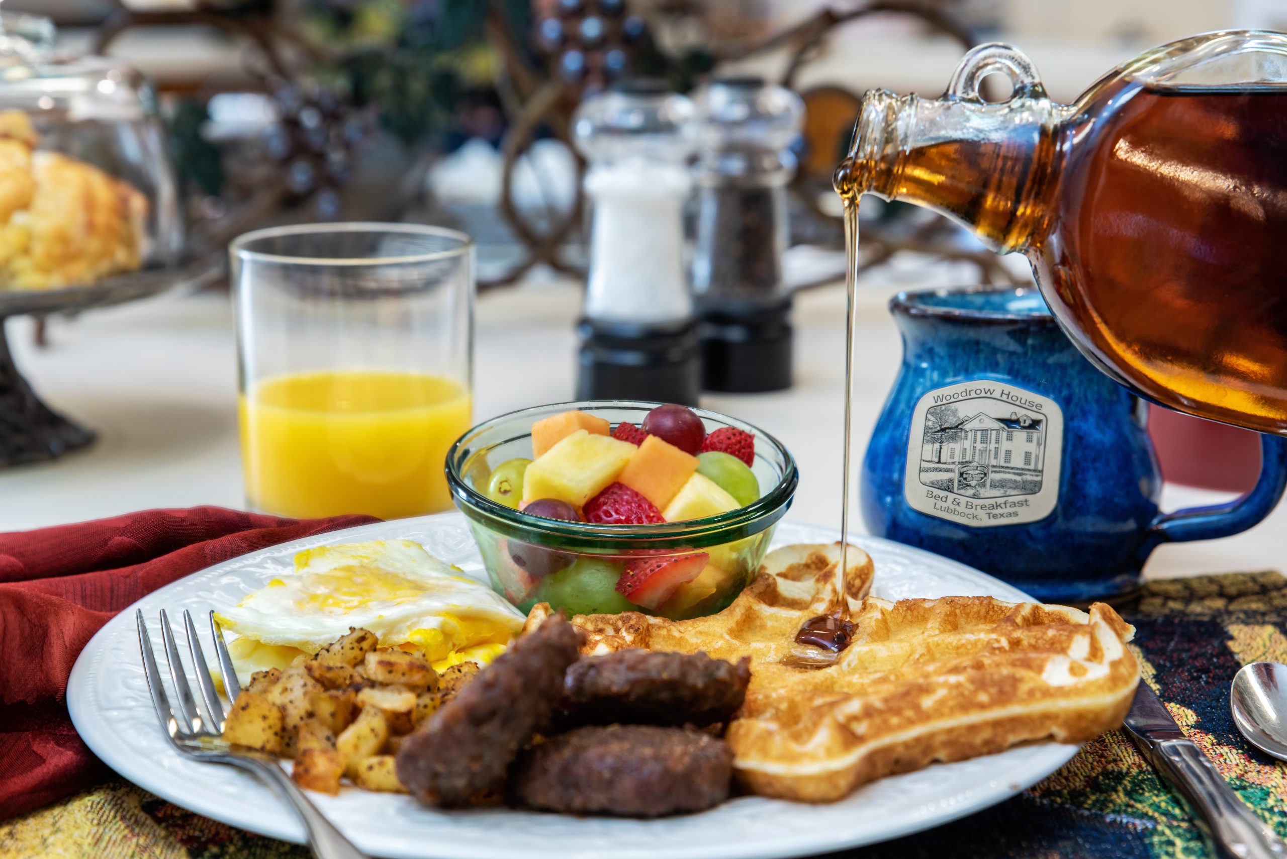 A delicious breakfast plate awaits at Woodrow House Bed & Breakfast, featuring sausage links, eggs, a waffle with syrup, potatoes, mixed fruit, orange juice, and coffee—all set on a table with a red napkin. Perfect Texas Tech lodging!.