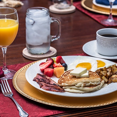 A hearty breakfast plate with pancakes, eggs, bacon, roasted potatoes, and fresh fruit awaits at Woodrow House Bed & Breakfast—a cozy B&B near Texas Tech University. Enjoy orange juice, coffee, and ice water for a perfect Texas Tech lodging experience.