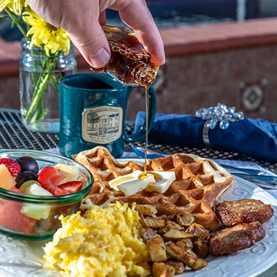 A hand pours syrup onto waffles topped with butter, alongside scrambled eggs, potatoes, sausage, and fruit—all part of a delicious breakfast at Woodrow House Bed & Breakfast, a welcoming B&B near Texas Tech University.