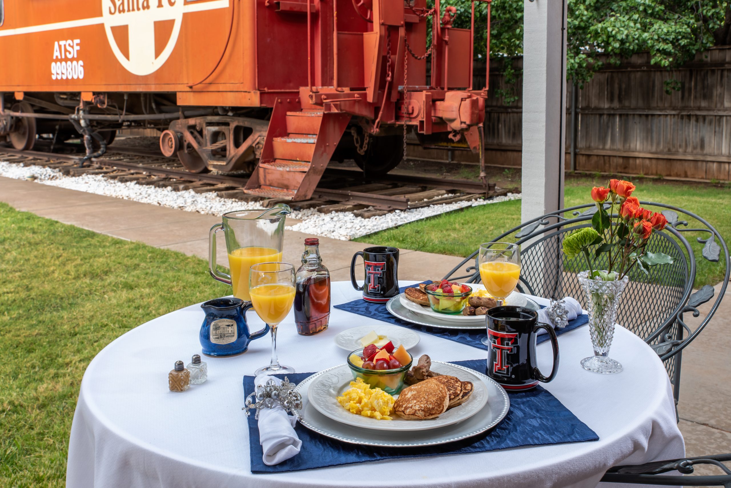 A round patio table set for breakfast with eggs, pancakes, fruit, and orange juice sits next to two chairs and a vase of orange flowers at Woodrow House Bed & Breakfast, a charming B&B near Texas Tech University. An orange caboose train car is on the track nearby.