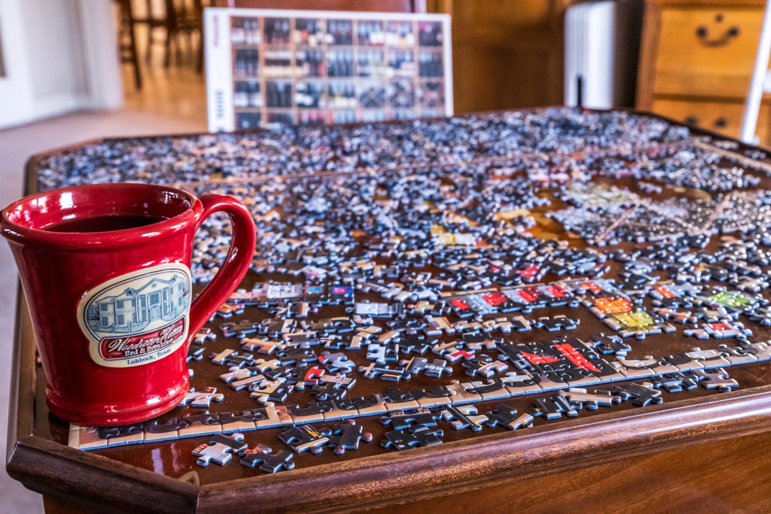 A red mug sits on the edge of a wooden table covered in scattered jigsaw puzzle pieces at Woodrow House Bed & Breakfast, with a partially completed puzzle and its box visible in the background.