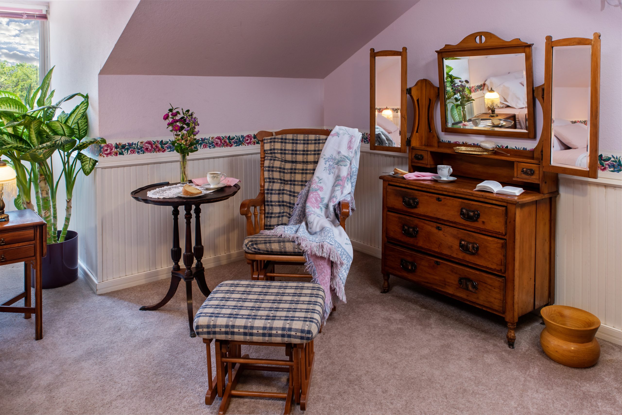 A cozy room at Woodrow House Bed & Breakfast features a cushioned rocking chair and footstool, round table with tea and flowers, wooden dresser with tri-fold mirror, potted plant, and pastel walls—ideal B&B near Texas Tech University.