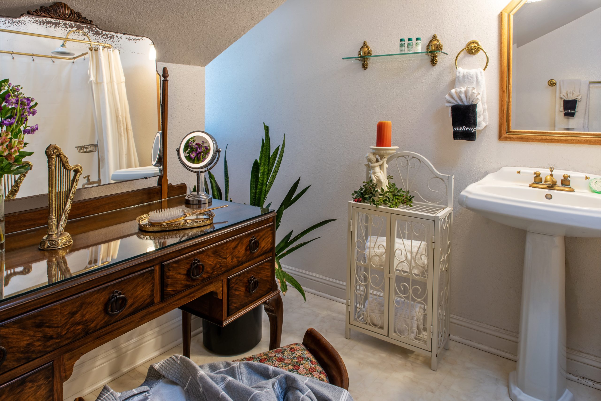 Elegant bathroom at Woodrow House Bed & Breakfast features a vintage wooden vanity, tabletop mirror, and fresh flowers. A decorative white shelf displays plants and towels—an inviting retreat for guests seeking B&B near Texas Tech University.