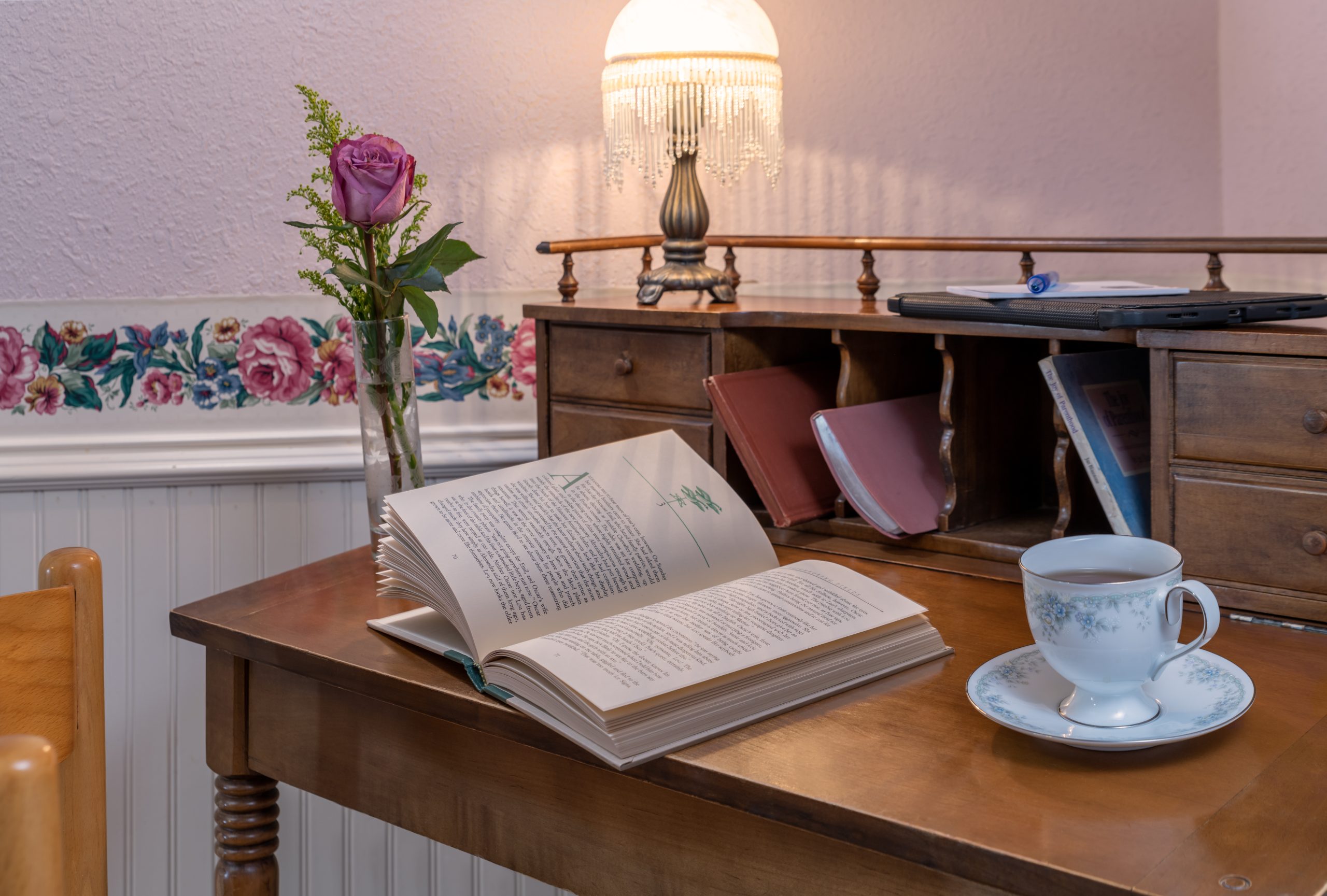 A vintage wooden desk with an open book, a teacup on a saucer, and a vase with a single purple rose sits beneath a beaded fringe lamp—evoking the charming ambiance of Woodrow House Bed & Breakfast, Texas Tech lodging at its finest.