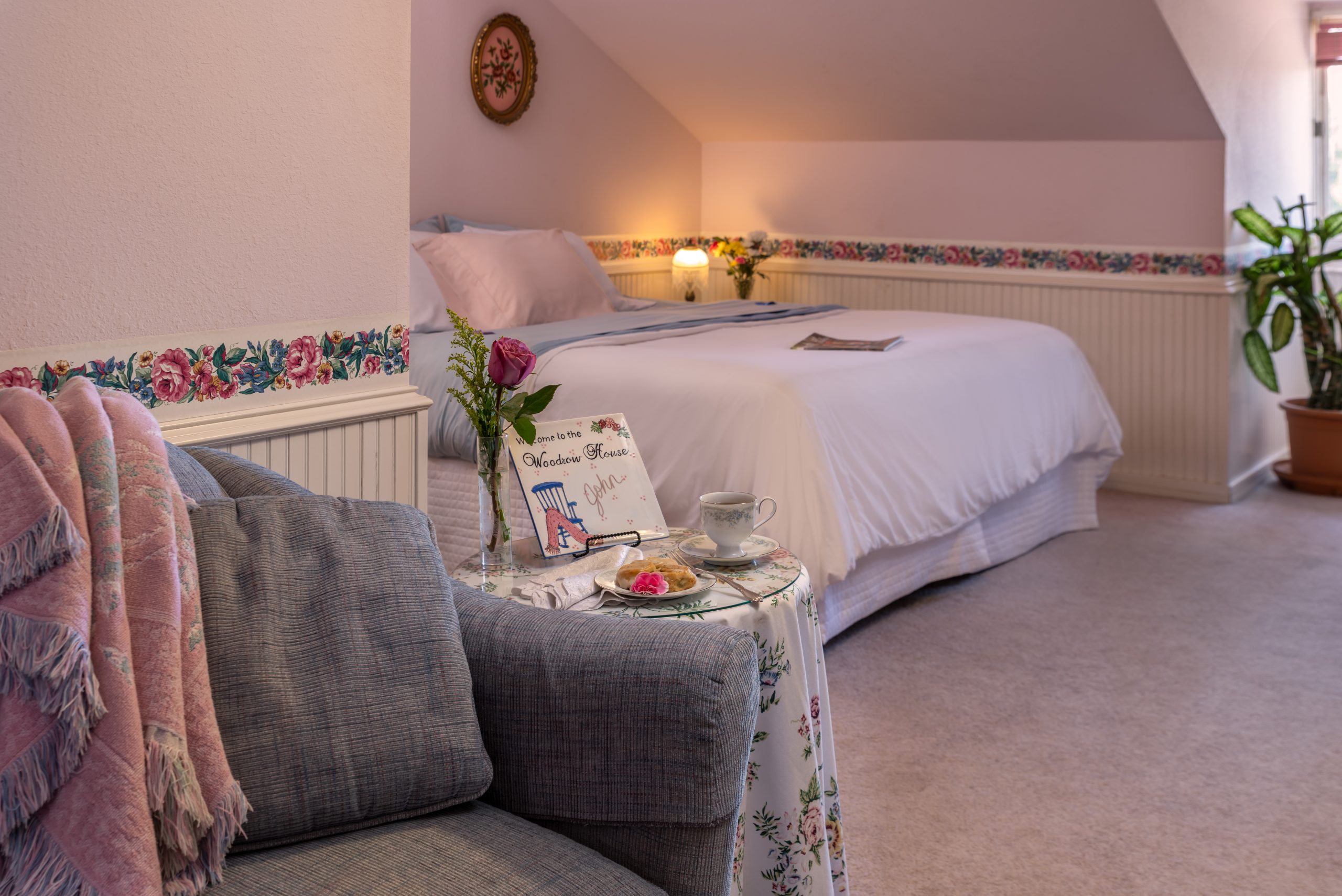 Cozy bedroom at Woodrow House Bed & Breakfast with a neatly made bed, floral wallpaper border, gray armchair with pink throw, and a small table set for tea. Sunlight filters in—a charming B&B near Texas Tech University.