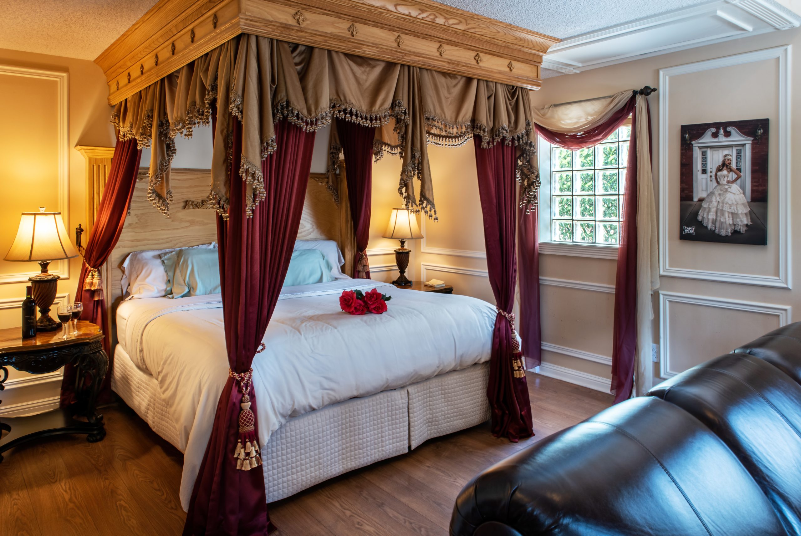 A luxurious bedroom at Woodrow House Bed & Breakfast features a grand canopy bed with gold and burgundy curtains, white bedding, red roses, elegant lamps, a leather sofa, and natural light—an ideal Texas Tech lodging choice.