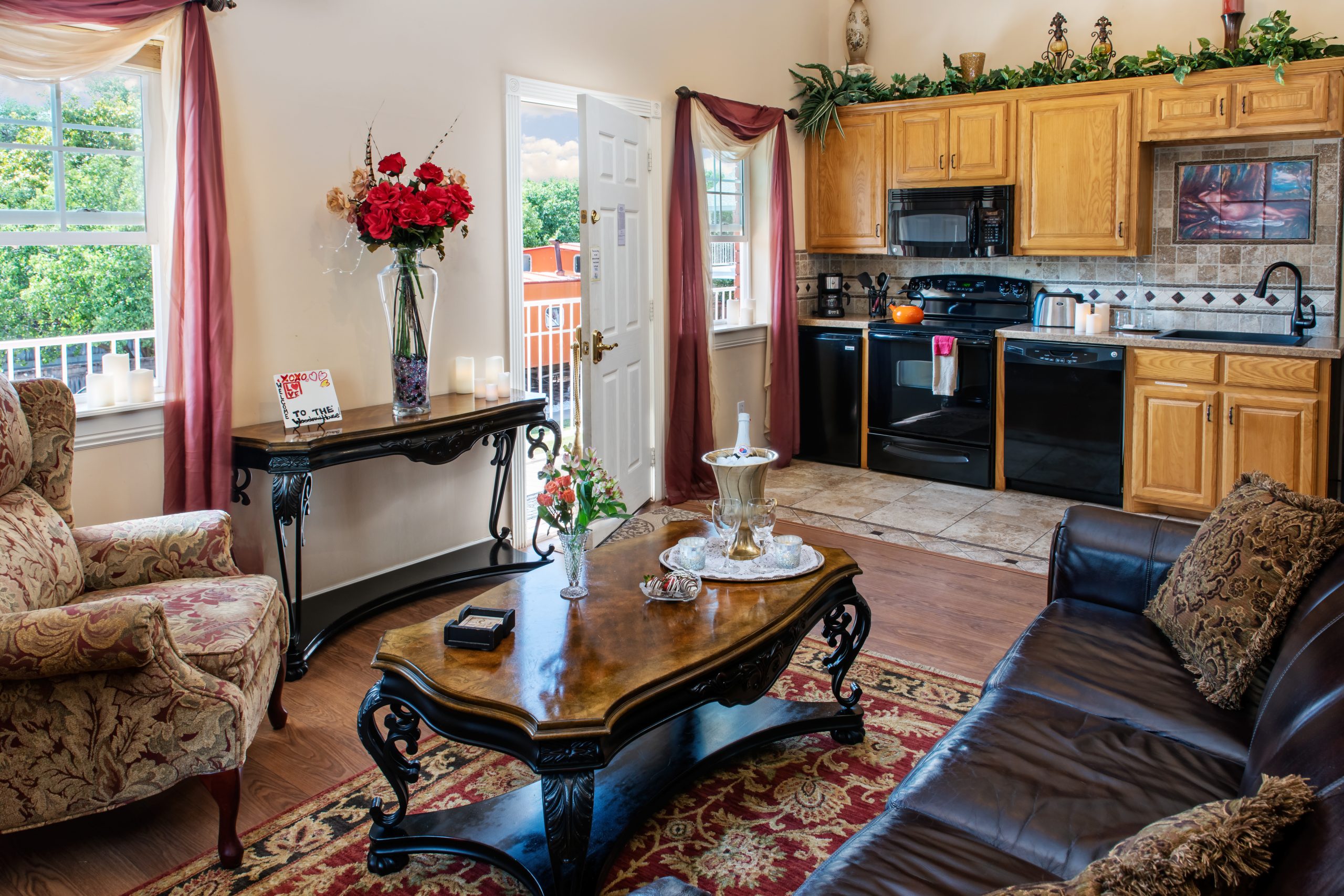 Cozy living room at Woodrow House Bed & Breakfast with a patterned armchair, dark leather sofa, ornate wood coffee table, and decorative console table. Open door reveals balcony view. Kitchen features wooden cabinets and black appliances. Ideal Texas Tech lodging.