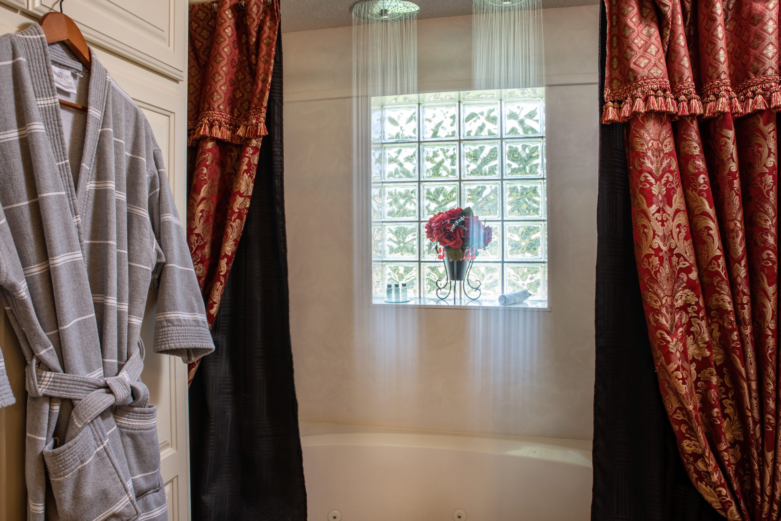 A bathtub with water flowing from a rainfall showerhead, flanked by red and gold patterned curtains, evokes comfort at this B&B near Texas Tech University. A window with glass blocks and a vase of red flowers sits above the tub; a gray bathrobe hangs nearby.