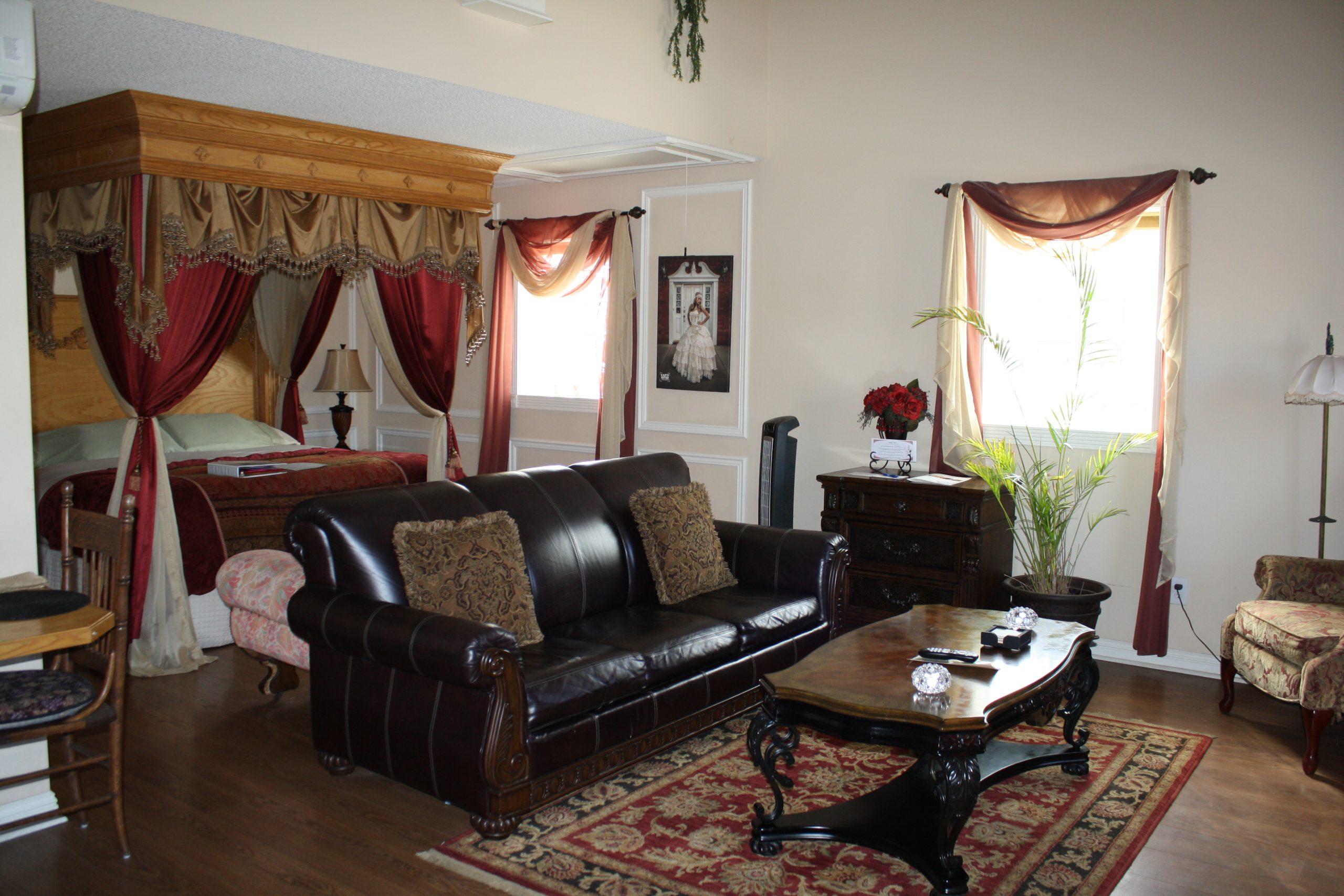 A cozy living area at Woodrow House Bed & Breakfast features a dark leather sofa, ornate coffee table, and armchair on a patterned rug. A canopied bed with red drapes sits in an alcove. Sunlight streams through windows with elegant curtains.
