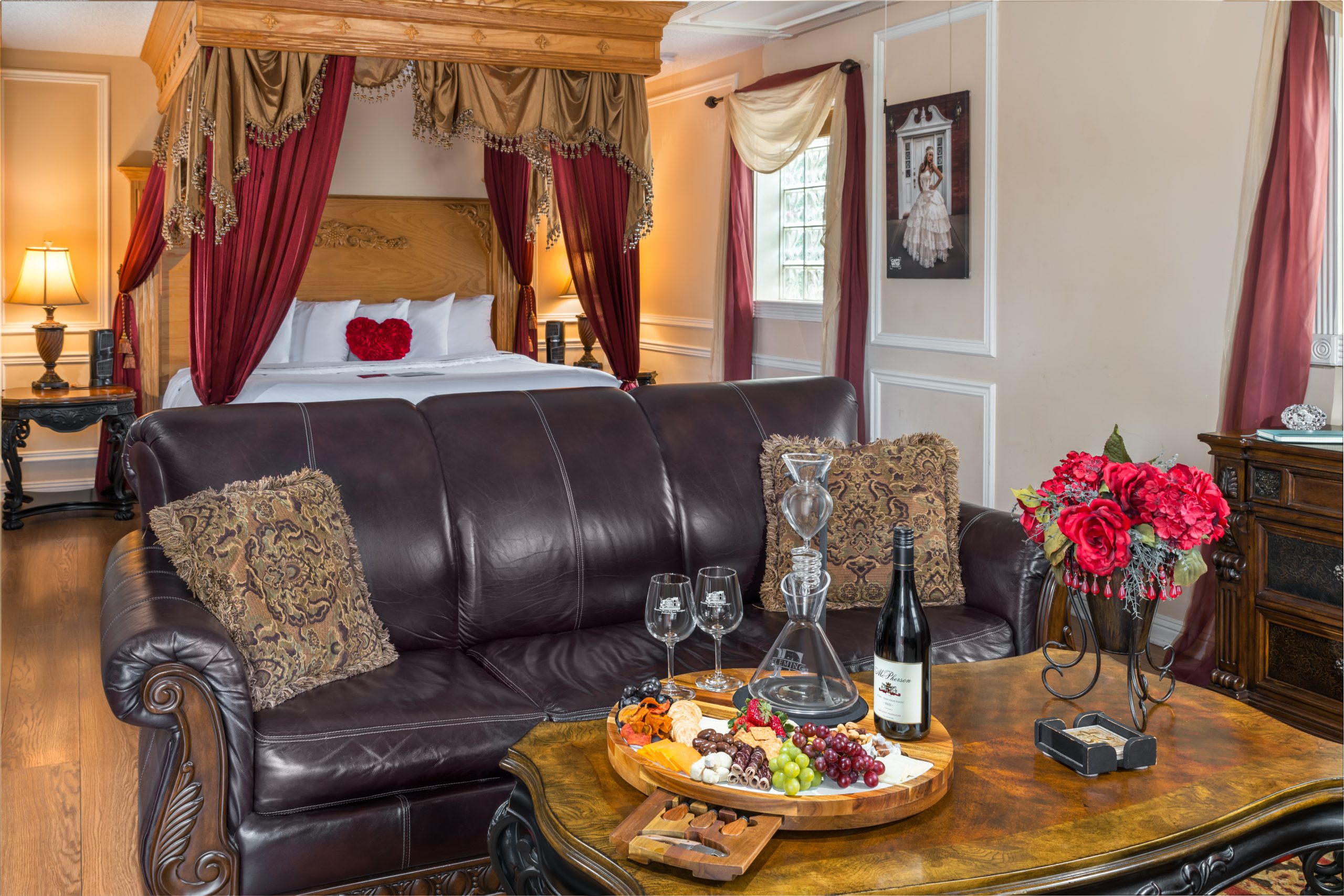 A luxurious bedroom at Woodrow House Bed & Breakfast near Texas Tech University features a canopy bed with red drapes, a leather sofa, and a wooden table set with wine, glasses, fruit, cheese, and flowers—all in an elegant, inviting atmosphere.