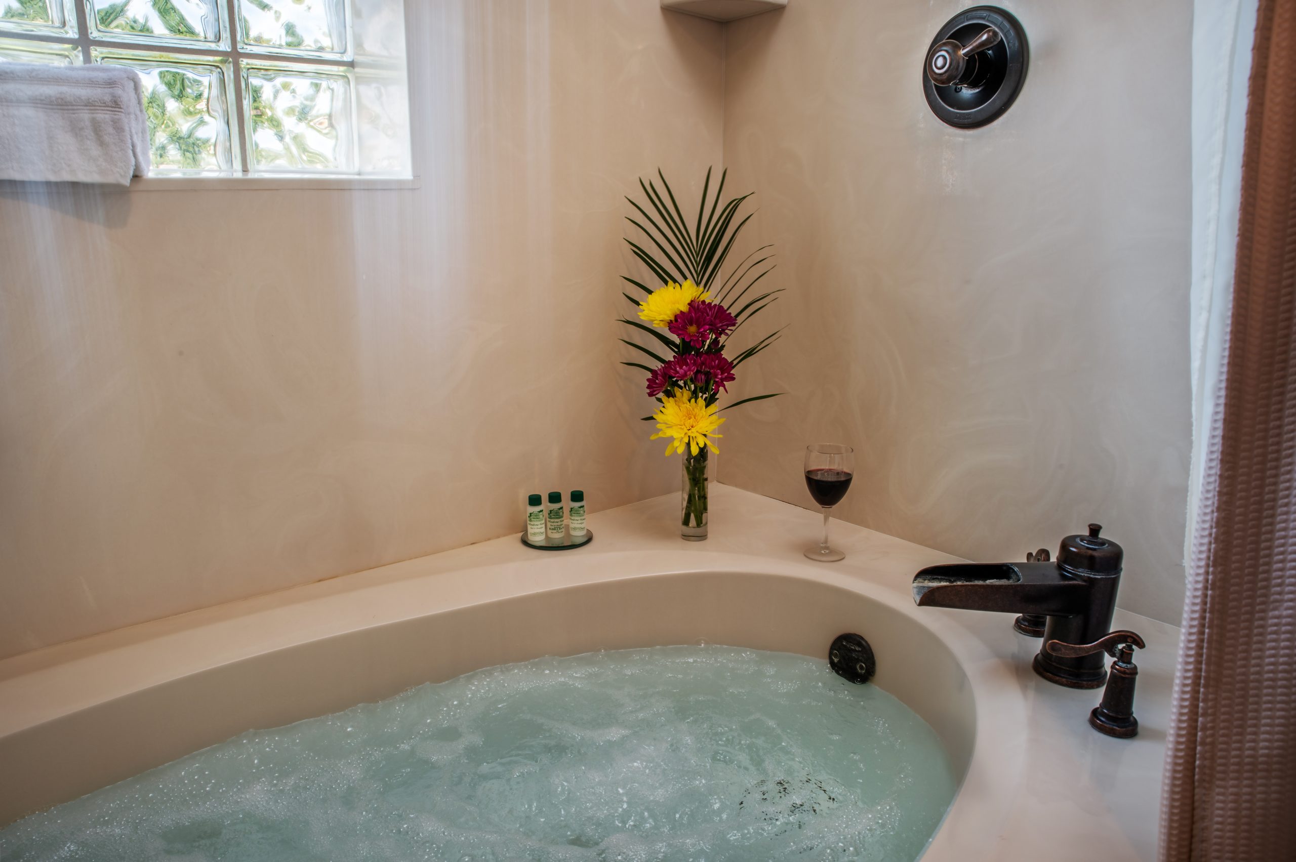 A filled bathtub with bubbling water sits beside a vase of colorful flowers, a glass of red wine, and small toiletries; a towel hangs by the glass block window at Woodrow House Bed & Breakfast, offering relaxing Texas Tech lodging.