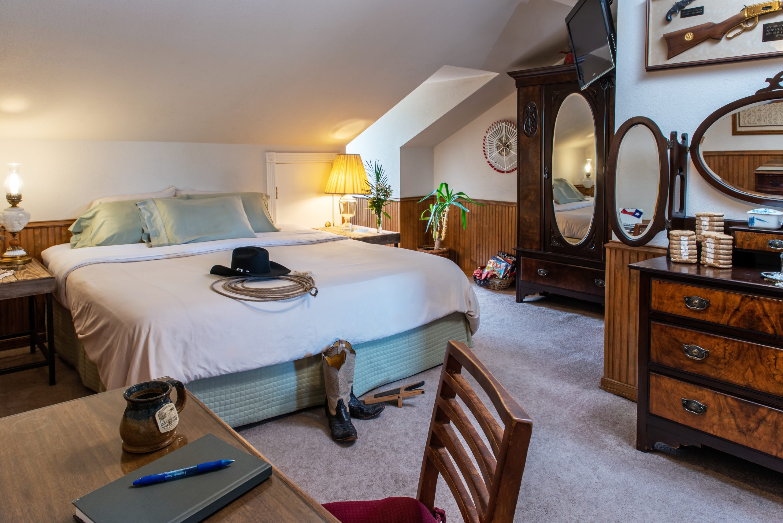A cozy bedroom at Woodrow House Bed & Breakfast features a large bed, cowboy hat, boots, and lasso on the floor, wooden furniture, mirror, dresser with coins, desk with notebook, and western decor—perfect Texas Tech lodging under a slanted ceiling.