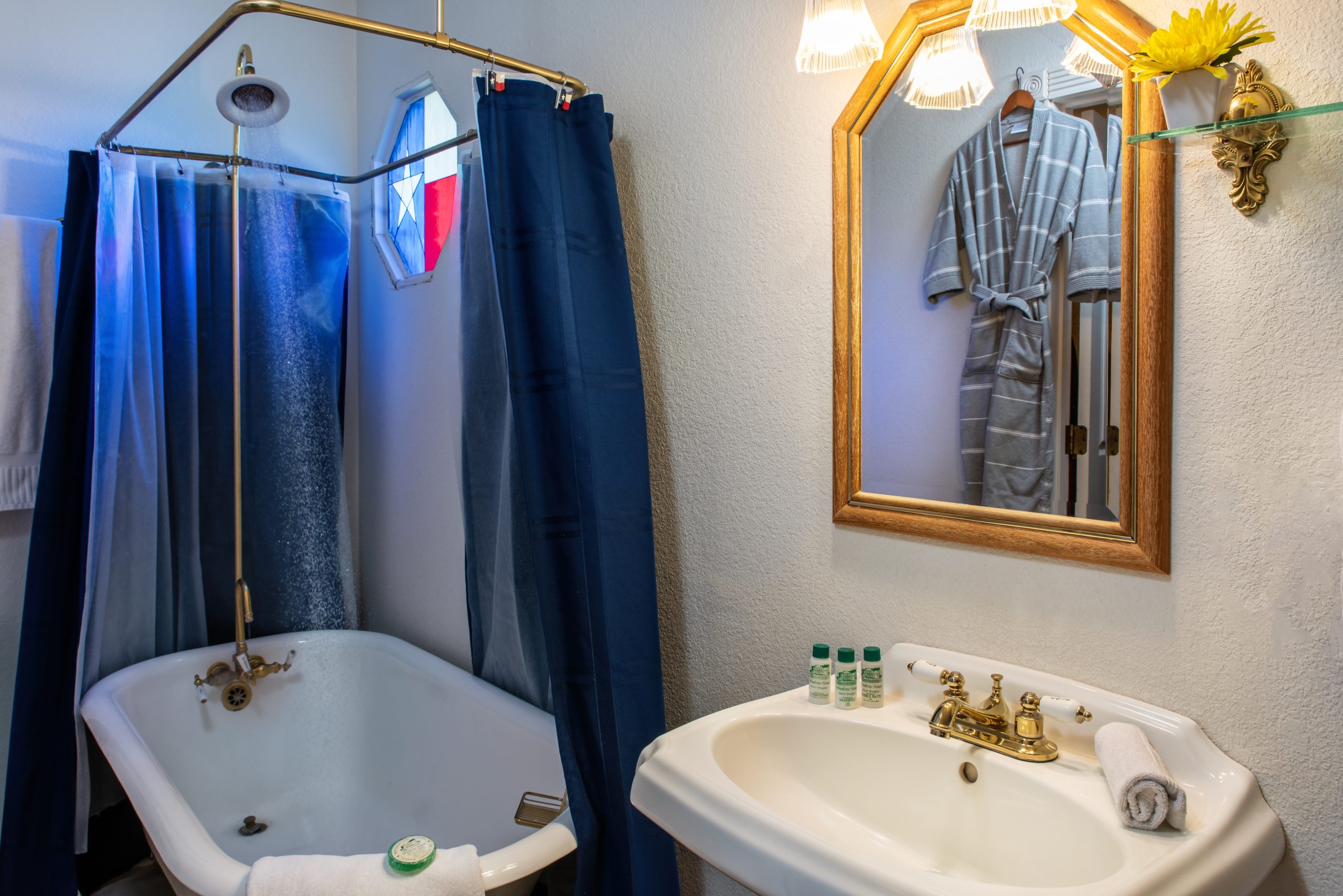 A bathroom at Woodrow House Bed & Breakfast features a white clawfoot tub with blue shower curtains, brass fixtures, a striped bathrobe reflected in the mirror, and a small shelf with a yellow flower—a cozy B&B near Texas Tech University.