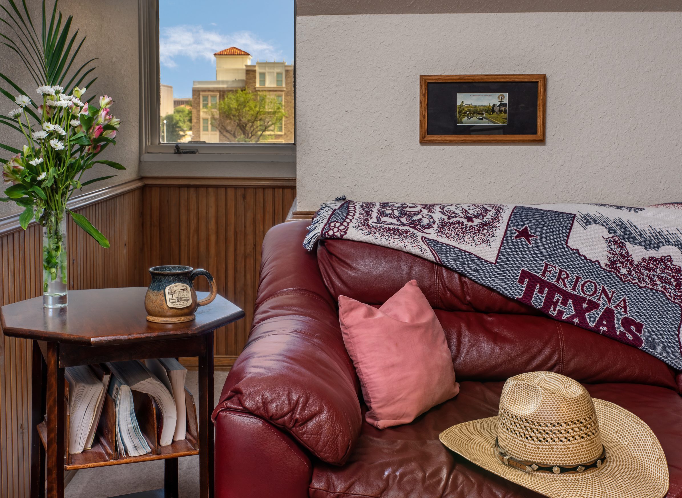 A cozy room at Woodrow House Bed & Breakfast features a red leather couch, pink pillow, straw hat, and Texas-themed throw. Nearby, a small table holds flowers and magazines—perfect Texas Tech lodging just steps from the university.