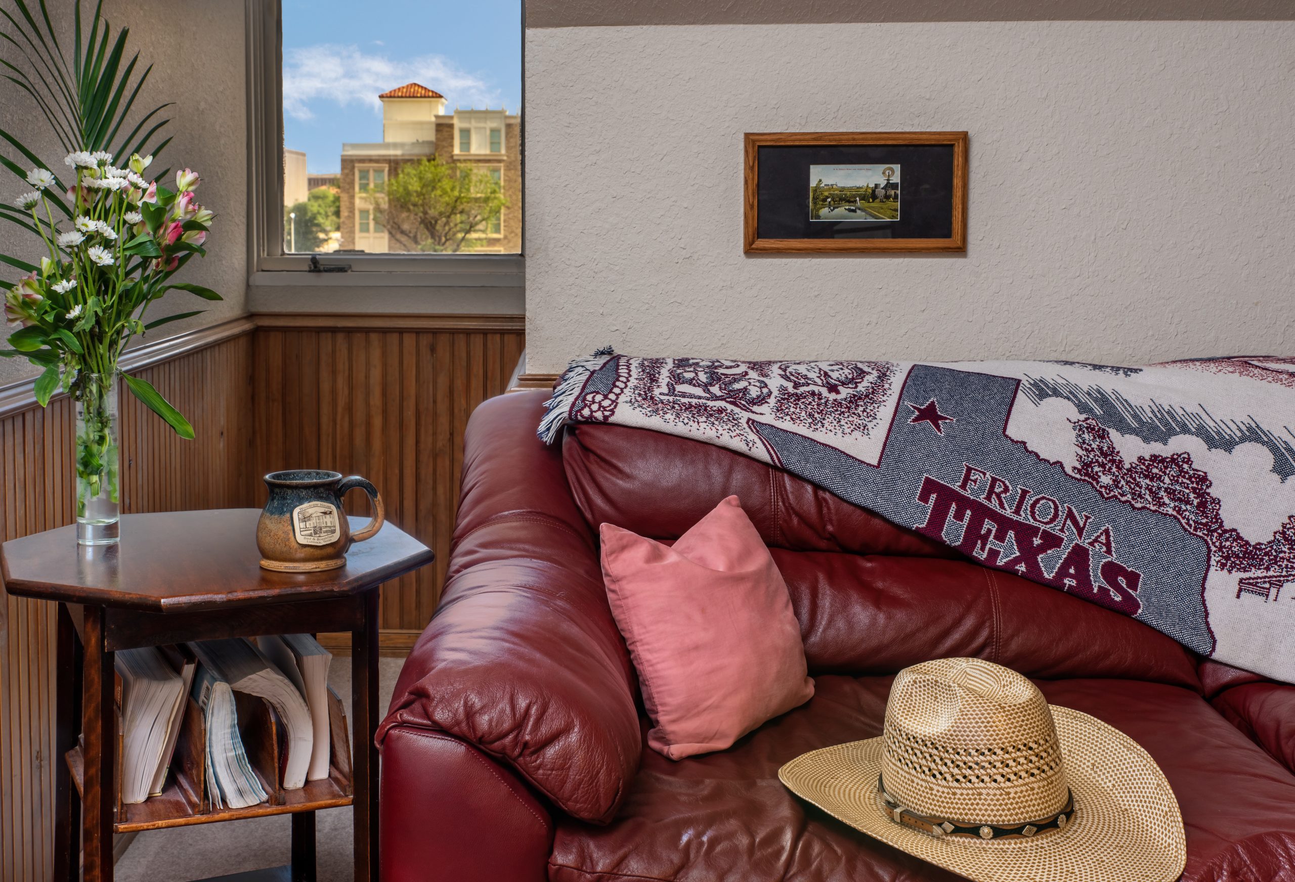 A cozy room at Woodrow House Bed & Breakfast features a red leather couch, pink pillow, straw hat, and Texas-themed throw. Nearby, a small table holds flowers and magazines—perfect Texas Tech lodging just steps from the university.