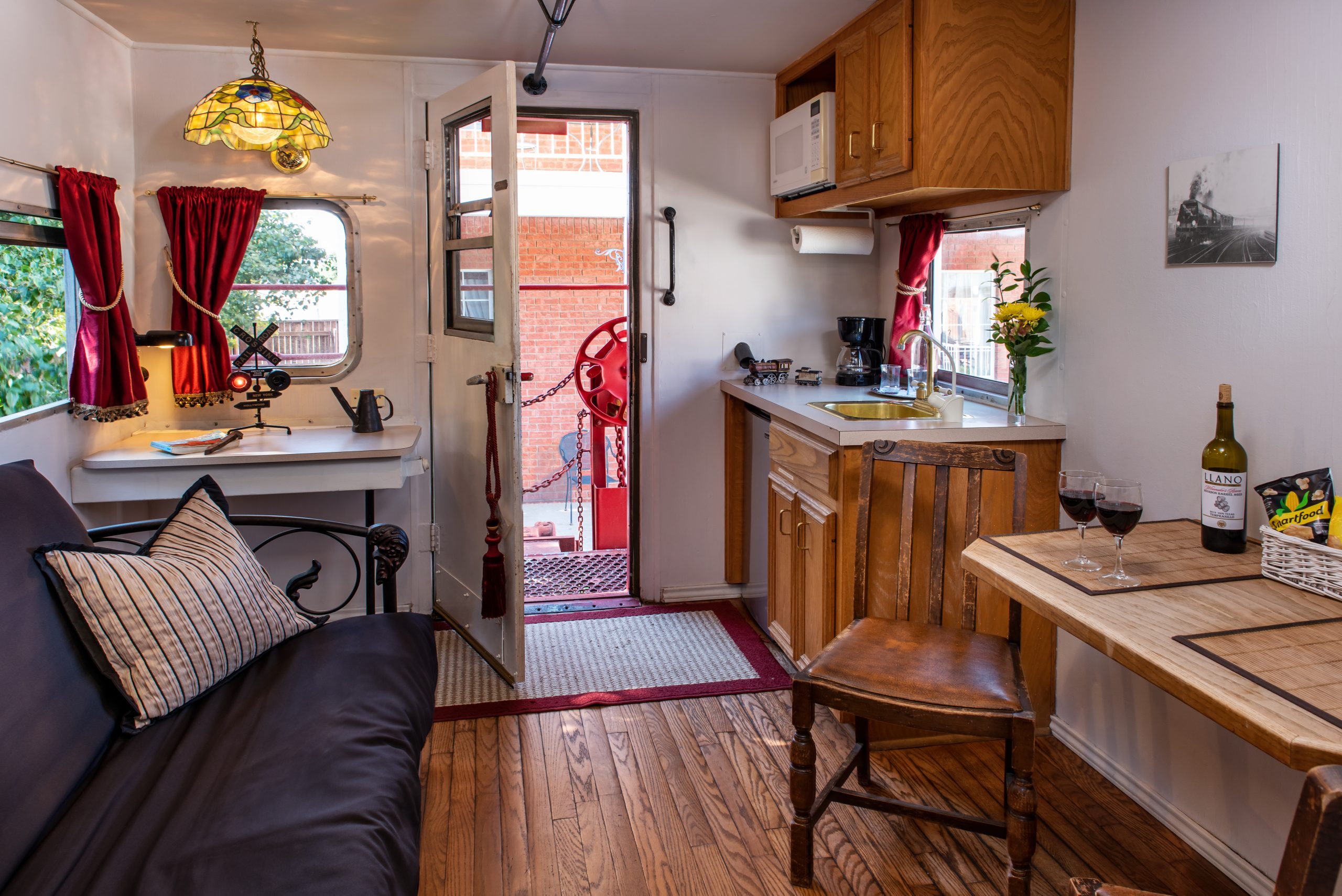 Cozy, well-lit camper interior with a small kitchen, wooden cabinets, table with wine, cushioned bench, and a door open to a patio. Red curtains, flowers, and a Tiffany-style lamp add charm to this B&B near Texas Tech University.