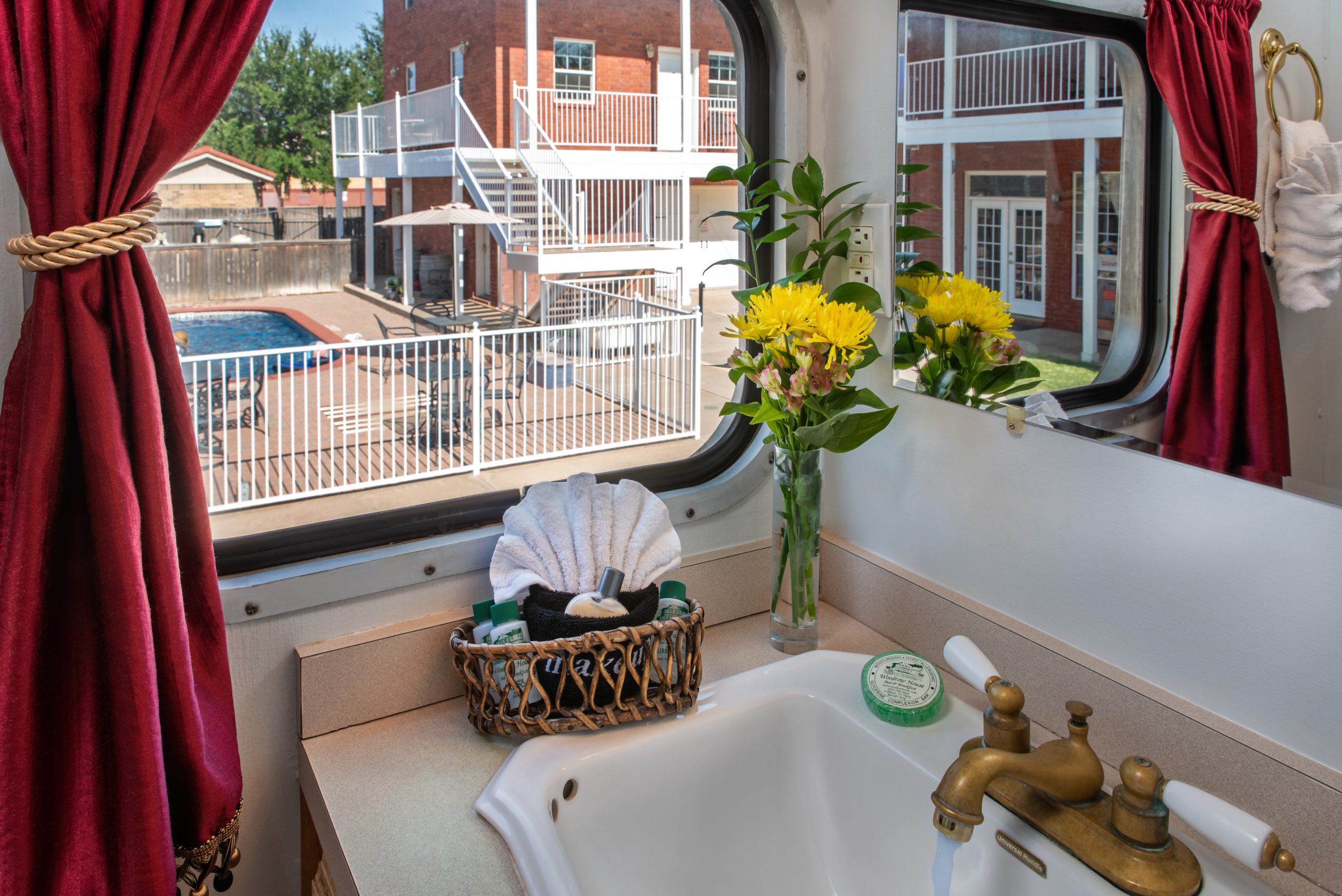 A bathroom sink with brass fixtures, a mirror, and a vase of yellow flowers. A basket with towels sits on the counter. Through the window at Woodrow House Bed & Breakfast, there’s a view of a pool and brick buildings with balconies.