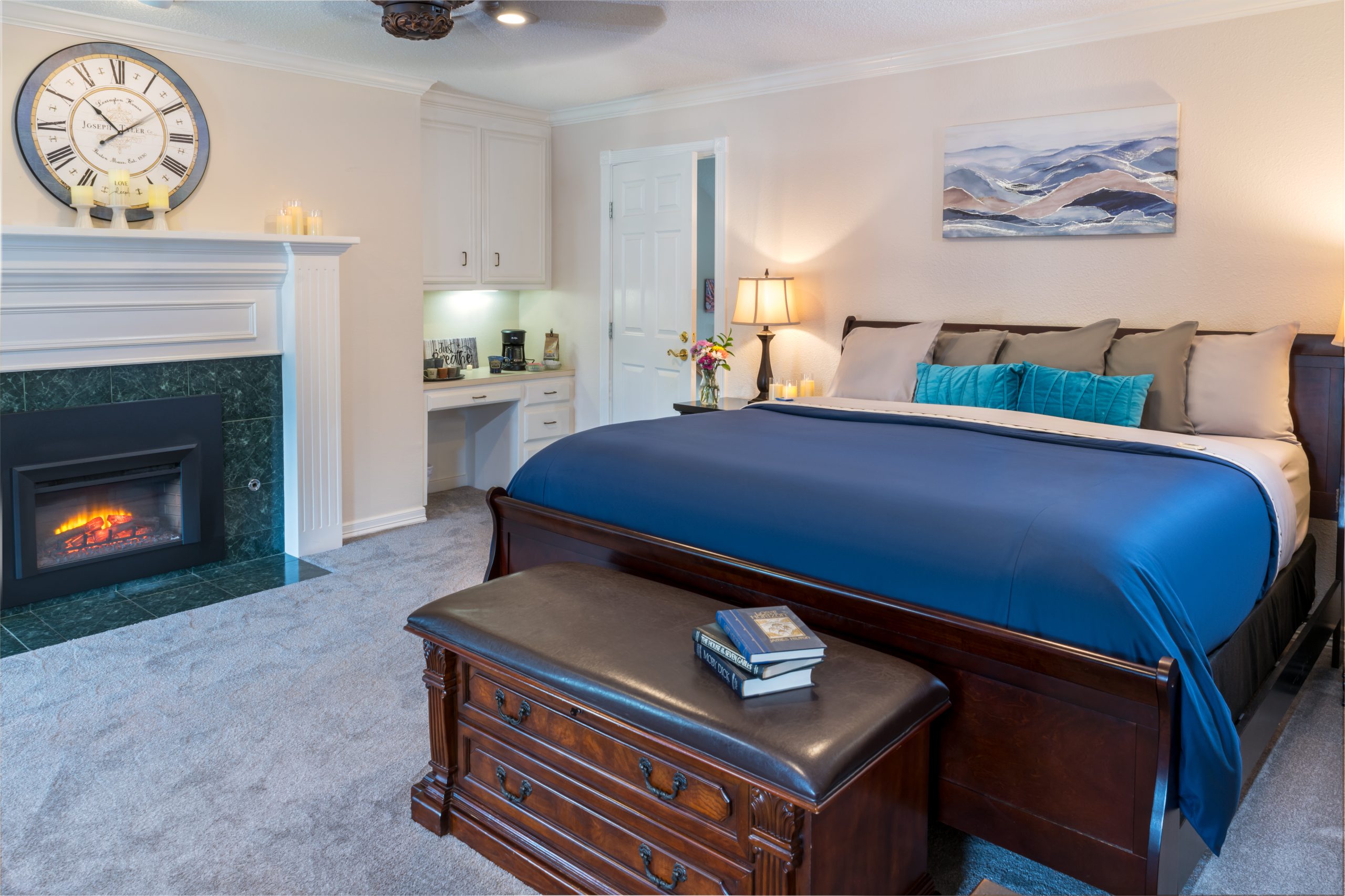 A cozy bedroom at Woodrow House Bed & Breakfast features a king-sized bed with blue and gray bedding, a fireplace, wall clock, desk with lamp, and a bench with books—perfect Texas Tech lodging just steps from the university.