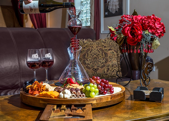 A wooden tray with assorted cheeses, fruits, and crackers sits on a table at Woodrow House Bed & Breakfast. Wine is being poured into a decanter, with two filled glasses nearby—a perfect Texas Tech lodging experience near the university.