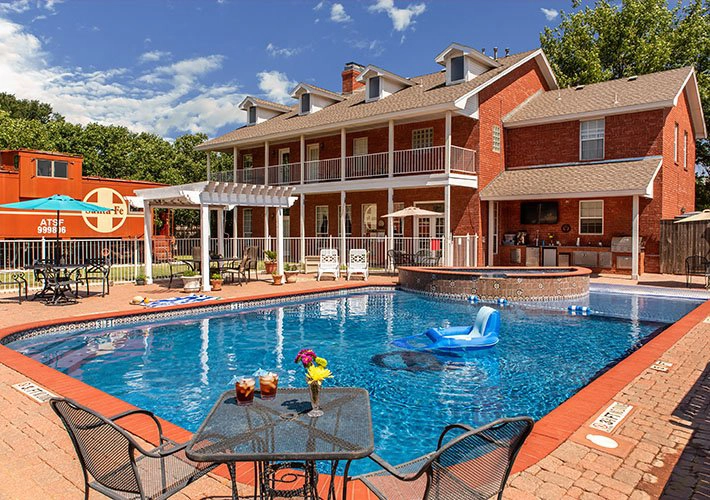 A backyard at Woodrow House Bed & Breakfast features a brick house, patio furniture, a swimming pool with floating lounge chair, hot tub, umbrellas, dining table with drinks and flowers—and even a large orange train car—ideal Texas Tech lodging.